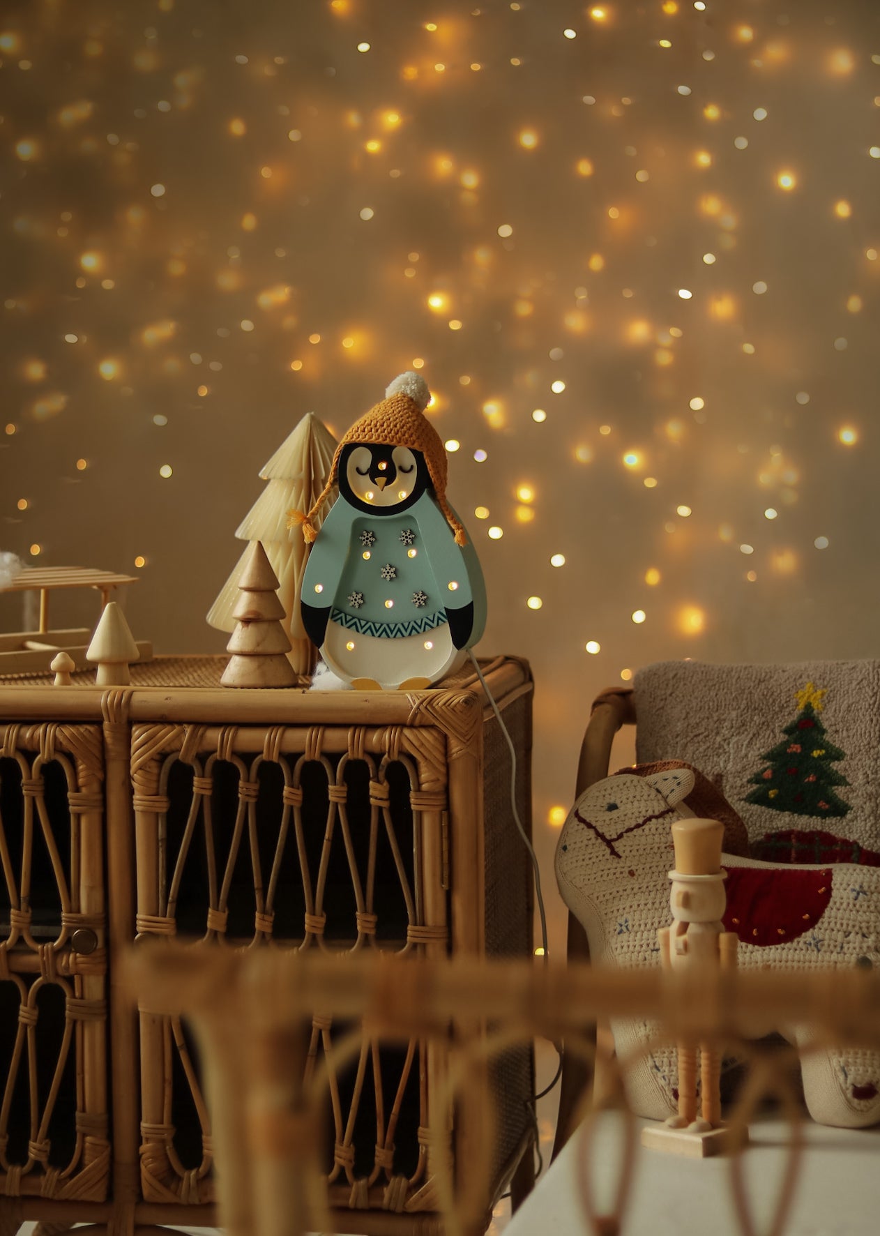 Eine festliche Szene mit warmen Weihnachtslichtern im Hintergrund. Im Vordergrund ein Korbschrank, dekoriert mit einem Little Lights – Baby Penguin Mini Christmas Edition | Blau von Little Lights und einem Miniatur-Weihnachtsbaum aus Holz. Neben dem Schrank steht ein gemütlicher Stuhl mit weihnachtlichen Kissen und sorgt für die perfekte Kinderzimmer-Dekoration.