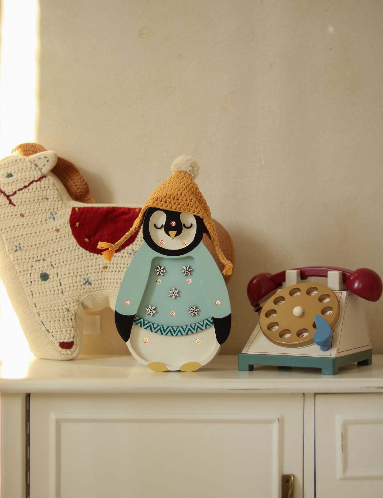 Ein bezauberndes Display mit einem ausgestopften, gehäkelten Pferdespielzeug, einer Little Lights - Baby Penguin Mini Christmas Edition | Blaue Figur mit Strickmütze und einem Spielzeugtelefon im Retro-Stil mit Wählscheibe. Sie sind alle auf einem weißen Schrank im Kinderzimmer-Dekorationsstil mit einer schlichten Wand im Hintergrund angeordnet.