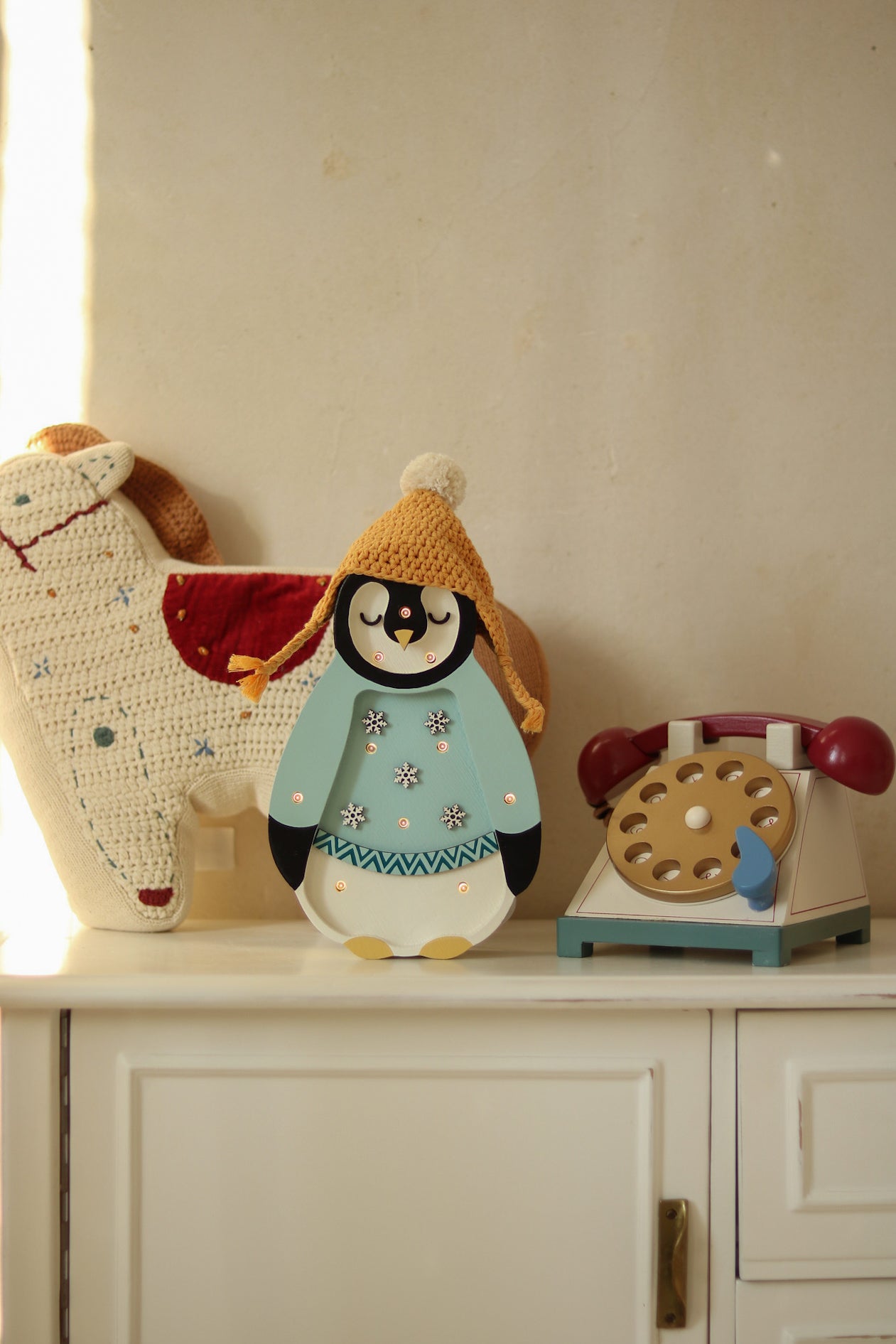 Ein bezauberndes Display mit einem ausgestopften, gehäkelten Pferdespielzeug, einer Little Lights - Baby Penguin Mini Christmas Edition | Blaue Figur mit Strickmütze und einem Spielzeugtelefon im Retro-Stil mit Wählscheibe. Sie sind alle auf einem weißen Schrank im Kinderzimmer-Dekorationsstil mit einer schlichten Wand im Hintergrund angeordnet.