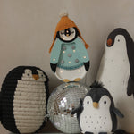 Eine Gruppe von Dekorationen mit Pinguinmotiven ist auf einer weißen Oberfläche angeordnet. Die Artikel umfassen einen Pinguin mit einer gestrickten orangefarbenen Mütze und einem blaugrünen Hemd, einen schwarz-weißen Plüschpinguin, ein Little Lights – Baby Penguin Mini Christmas Edition | Blau, perfekt für die Kinderzimmer-Dekoration, eine große Pinguinfigur und eine glänzende Discokugel in der Mitte.
