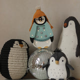 Eine Gruppe von Dekorationen mit Pinguinmotiven ist auf einer weißen Oberfläche angeordnet. Die Artikel umfassen einen Pinguin mit einer gestrickten orangefarbenen Mütze und einem blaugrünen Hemd, einen schwarz-weißen Plüschpinguin, ein Little Lights – Baby Penguin Mini Christmas Edition | Blau, perfekt für die Kinderzimmer-Dekoration, eine große Pinguinfigur und eine glänzende Discokugel in der Mitte.
