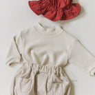 Kinderoutfit bestehend aus einem cremefarbenen Langarmshirt aus hautfreundlichem Material, hellbeiger Hose mit Gummibund und Taschen sowie einer roten Mütze mit dezentem Muster. Die Teile sind übersichtlich auf einem einfarbigen Untergrund angeordnet, mit dem Tube Tee - Bequemes und Stilvolles T-Shirt für die Kleinsten von Bebe Holic.