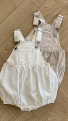 Zwei Bebe Holic Leinen-Latzhosen mit Schnallen, eine in Off-White und die andere in Beige, liegen auf einem hellen Holzboden. Jeder Strampler aus atmungsaktiver Baumwolle hat verstellbare Träger mit Metallschnallen und eine Vordertasche mit gerafftem Stoff um die Taille und Beinöffnungen.