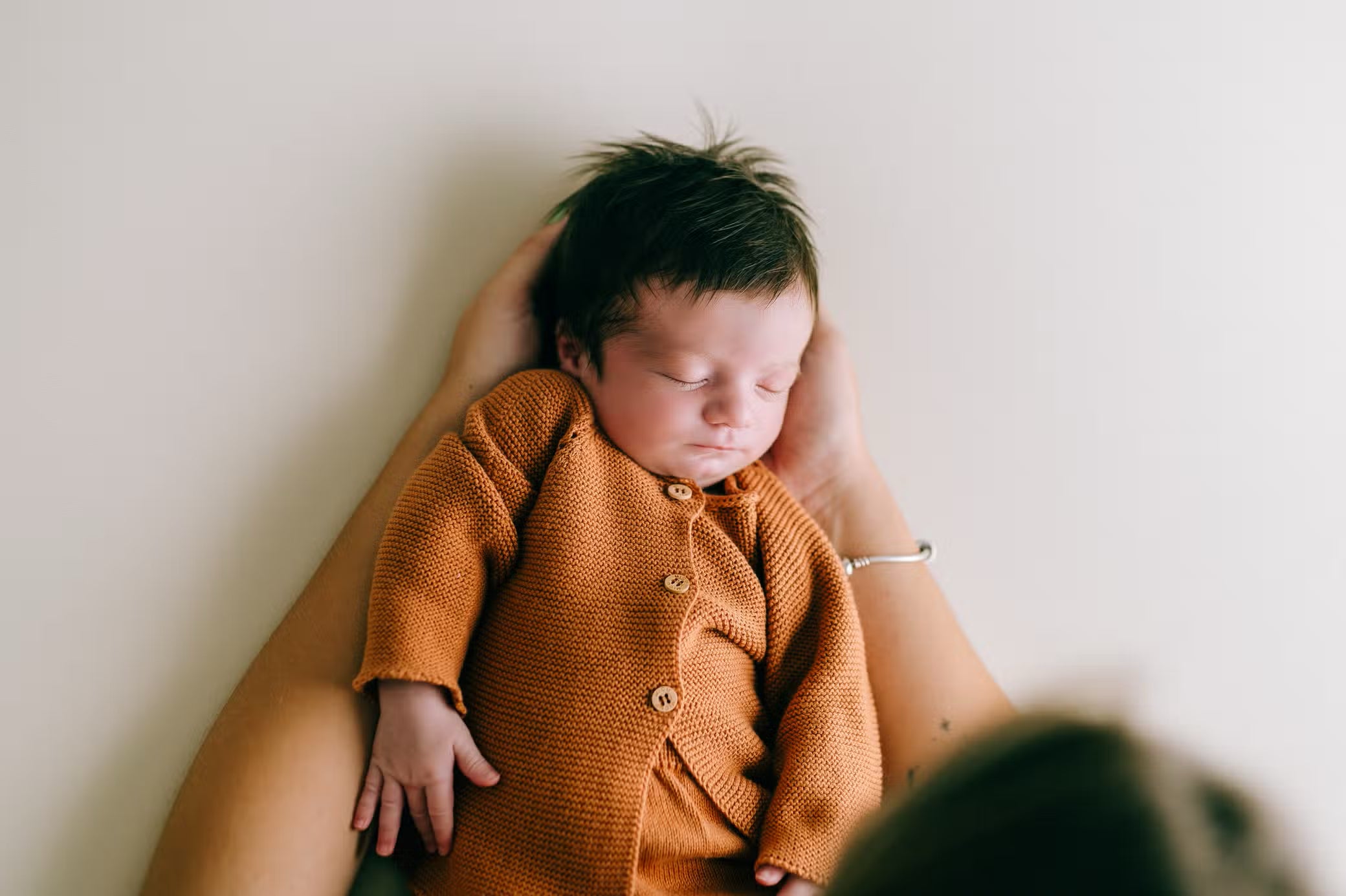 Ein friedliches Baby mit dunklem Haar schläft tief und fest, während es ein Micu Micu Newborn Pack ziegelrot 3-6m trägt. Das Neugeborene wird sanft in einem Paar erwachsener Hände vor einem weichen, hellen Hintergrund gewiegt.