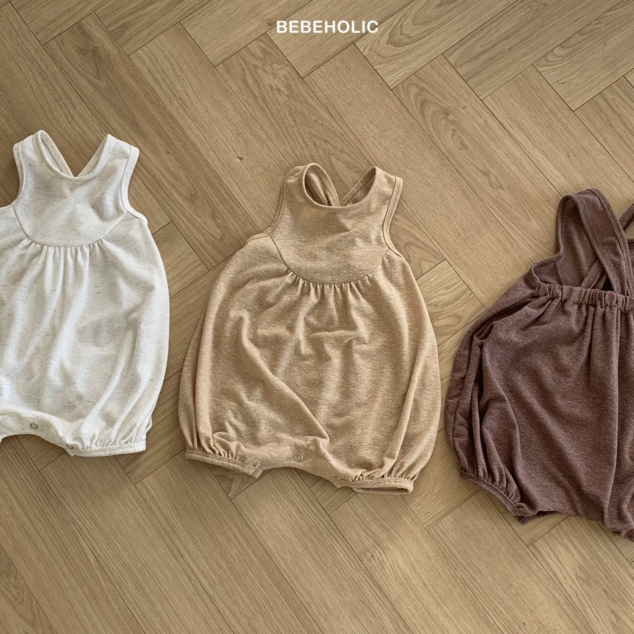 Drei Bebe Holic Leinen-Bodys in neutralen Farbtönen (Off-White, Beige und Braun) liegen auf einem hölzernen Fischgrätboden. Die Strampler haben ein einfaches, ärmelloses Design mit elastischen Beinöffnungen für zusätzlichen Komfort für Kleinkinder. Der Text „BEBE HOLIC“ steht oben in der Mitte des Bildes.