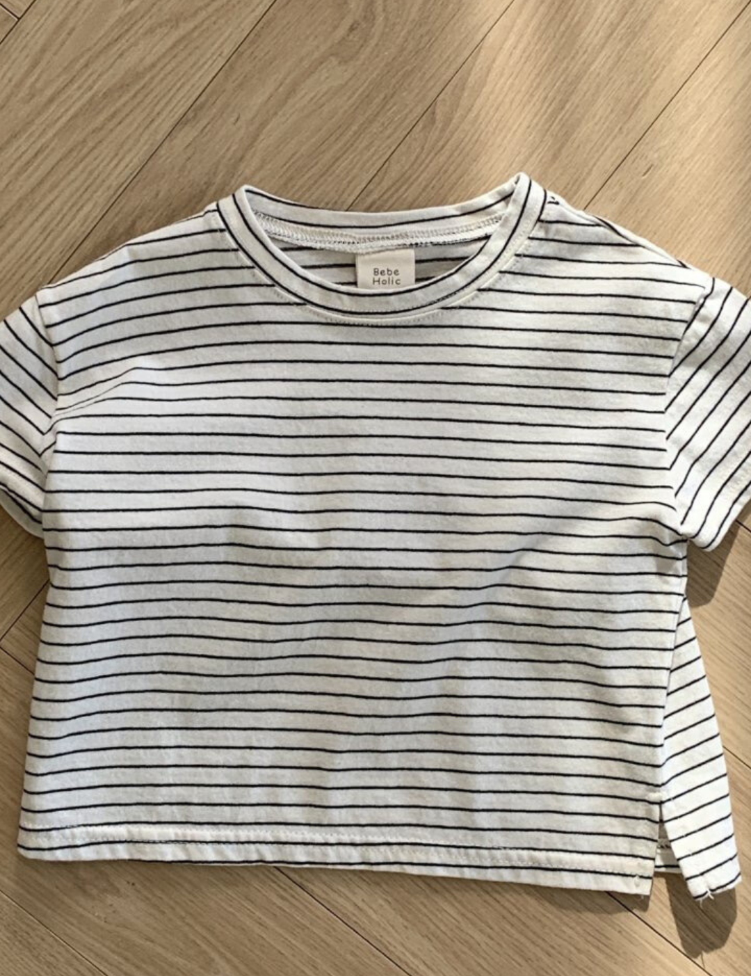 Ein kurzärmliges, weißes Crop-Top mit schwarzen Querstreifen liegt flach auf einem Holzboden. Dieses Basic ST Tee von Bebe Holic hat einen runden Ausschnitt und ein Etikett im Kragen. Der Holzboden mit seinem hellen, natürlichen Finish und Fischgrätenmuster unterstreicht seinen klassischen Stil und bequemen Tragekomfort.