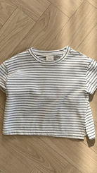 Ein kurzärmliges, weißes Crop-Top mit schwarzen Querstreifen liegt flach auf einem Holzboden. Dieses Basic ST Tee von Bebe Holic hat einen runden Ausschnitt und ein Etikett im Kragen. Der Holzboden mit seinem hellen, natürlichen Finish und Fischgrätenmuster unterstreicht seinen klassischen Stil und bequemen Tragekomfort.