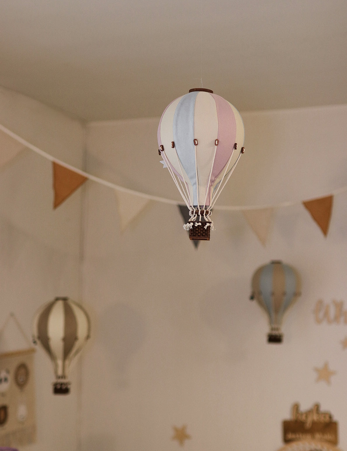 Ein mit vielseitiger Dekoration dekorierter Raum mit hängenden Ornamenten in Form von Heißluftballons. Im Vordergrund ist ein blau-rosa gestreifter dekorativer Ballon – Beige, Mint, Grau und Lila | Einzigartiges Heißluftballon-Ornament für Ihr Zuhause von Superballoon zu sehen, im Hintergrund sind weitere dekorative Ballons in gedeckten Farben zu sehen. Ein dreieckiges Wimpelbanner hängt anmutig quer durch den Raum.