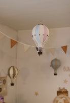 Ein mit vielseitiger Dekoration dekorierter Raum mit hängenden Ornamenten in Form von Heißluftballons. Im Vordergrund ist ein blau-rosa gestreifter dekorativer Ballon – Beige, Mint, Grau und Lila | Einzigartiges Heißluftballon-Ornament für Ihr Zuhause von Superballoon zu sehen, im Hintergrund sind weitere dekorative Ballons in gedeckten Farben zu sehen. Ein dreieckiges Wimpelbanner hängt anmutig quer durch den Raum.