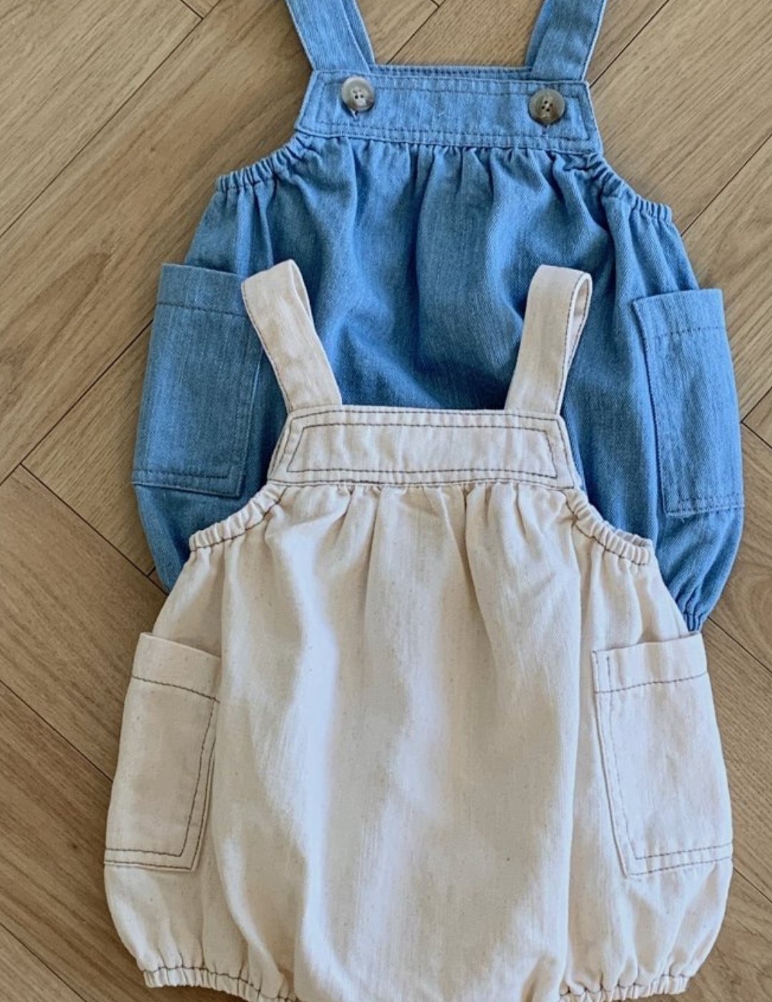 Zwei Bebe Holic Denim Pocket Body Suits sind auf einem Holzboden mit Fischgrätenmuster ausgelegt. Der obere Body Suit ist Jeansblau mit großen Vordertaschen und verstellbaren Trägern, die zusätzliche Bewegungsfreiheit bieten. Der untere Body Suit ist Beige mit passenden großen Vordertaschen und ähnlichem Trägerdesign.