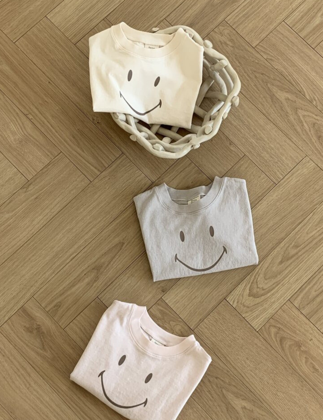 Drei gefaltete Bebe Holic Smile Tees aus weicher Baumwolle mit Smileys darauf sind auf einem Holzboden angeordnet. Zwei T-Shirts liegen direkt auf dem Boden, während eines in einem geflochtenen Korb liegt. Die T-Shirts sind in hellen Creme-, Grau- und Rosatönen gehalten.