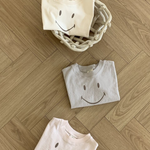 Drei gefaltete Bebe Holic Smile Tees aus weicher Baumwolle mit Smileys darauf sind auf einem Holzboden angeordnet. Zwei T-Shirts liegen direkt auf dem Boden, während eines in einem geflochtenen Korb liegt. Die T-Shirts sind in hellen Creme-, Grau- und Rosatönen gehalten.