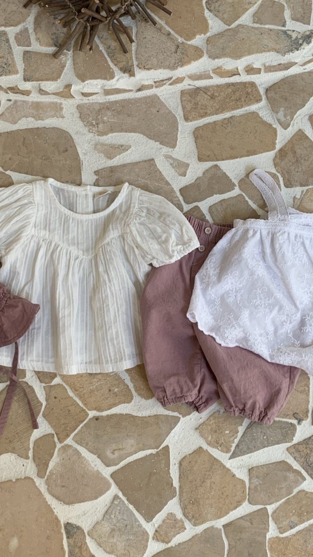 Babys Bebe Holic Raoul-Bluse auf rustikalem Steinboden, bestehend aus einem weißen Rüschenoberteil mit luftiger Ärmelgestaltung und einem staubrosafarbenen Overall mit Spitzendetails.