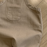 Ein hellbrauner Bebe Holic Twill-Body mit verstellbarem Träger und einer kleinen Brusttasche. Der Strampler aus weicher Baumwolle hat Druckknöpfe an der Unterseite zum einfachen Windelwechseln. Er liegt flach auf einem Holzboden, wobei Licht und Schatten auf der Oberfläche sichtbar sind.