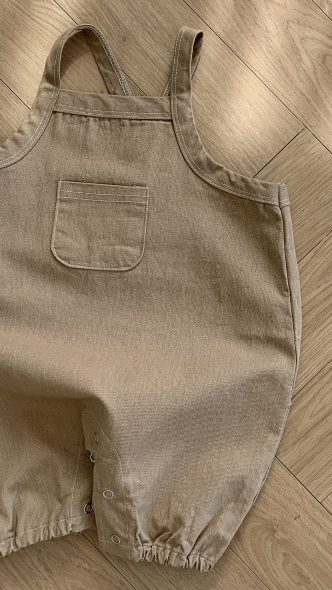 Ein hellbrauner Bebe Holic Twill-Body mit verstellbarem Träger und einer kleinen Brusttasche. Der Strampler aus weicher Baumwolle hat Druckknöpfe an der Unterseite zum einfachen Windelwechseln. Er liegt flach auf einem Holzboden, wobei Licht und Schatten auf der Oberfläche sichtbar sind.