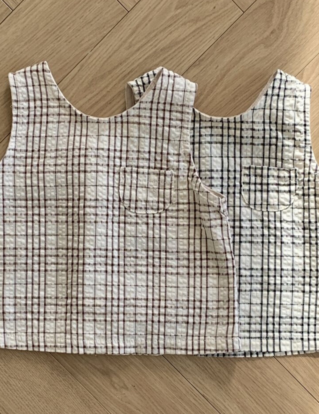 Zwei Summer Check Sleeveless Tees von Bebe Holic aus strukturiertem Stoff mit Gittermuster liegen auf einem Holzboden. Ein Oberteil ist weiß mit rotem Gitter, das andere weiß mit schwarzem Gitter. Jedes Oberteil hat eine kleine Tasche auf der Brust, die einen Hauch modischen Flairs verleiht und perfekt in jede Bebeholic-Kollektion passt.