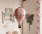 Ein skurriles Kinderzimmer verfügt über einen hängenden Superballoon-Dekoballon in BEIGE/PUDERROSA. Der Hintergrund besteht aus gerahmten Kunstwerken, einem grauen Schmetterlings-Wandornament und einer Reihe sternförmiger Girlanden, die dem Raum eine verträumte und gemütliche Atmosphäre verleihen.