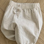 Ein Paar beige Bebe Holic Natural Pants mit elastischer Taille und zwei Knöpfen auf der Vorderseite, flach auf einem hellen Holzboden mit Fischgrätenmuster. Diese vielseitigen Must-have-Shorts sind ein Muss für die Garderobe Ihres Babys.