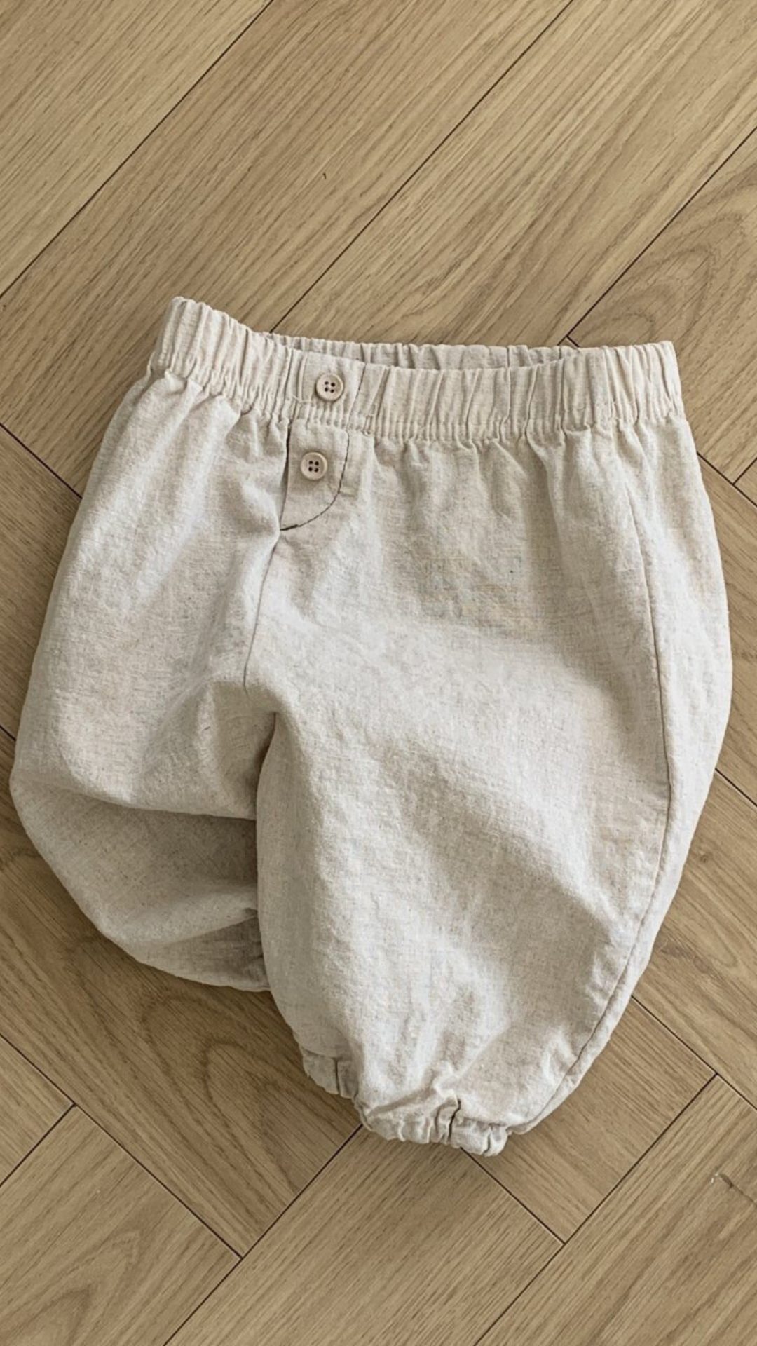 Ein Paar beige Bebe Holic Natural Pants mit elastischer Taille und zwei Knöpfen auf der Vorderseite, flach auf einem hellen Holzboden mit Fischgrätenmuster. Diese vielseitigen Must-have-Shorts sind ein Muss für die Garderobe Ihres Babys.