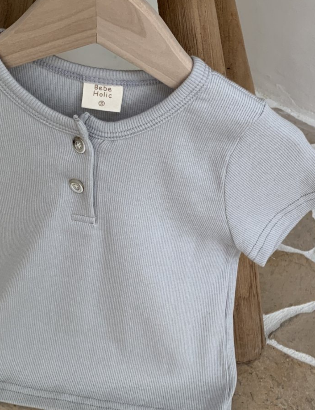 Ein hellgraues, geripptes Baby-Kurzarmshirt mit zwei Knöpfen am Kragen hängt auf einem hölzernen Kleiderbügel. Das ST-T-Shirt aus atmungsaktiver Baumwolle liegt vor einem Holzhocker auf einem Steinfliesenboden. Auf dem Etikett im Inneren steht „Bebe Holic“.