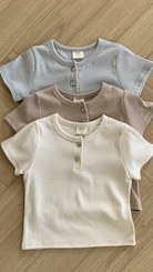 Drei kurzärmelige Kleinkindershirts im Henley-Stil von Bebe Holic ST Tee in Pastellfarben (Hellblau, Hellbraun und Cremeweiß) sind ordentlich auf einer Holzoberfläche angeordnet. Jedes Shirt hat eine gerippte Textur, atmungsaktives Baumwollmaterial und zwei Knöpfe an den Knopfleisten am Kragen.