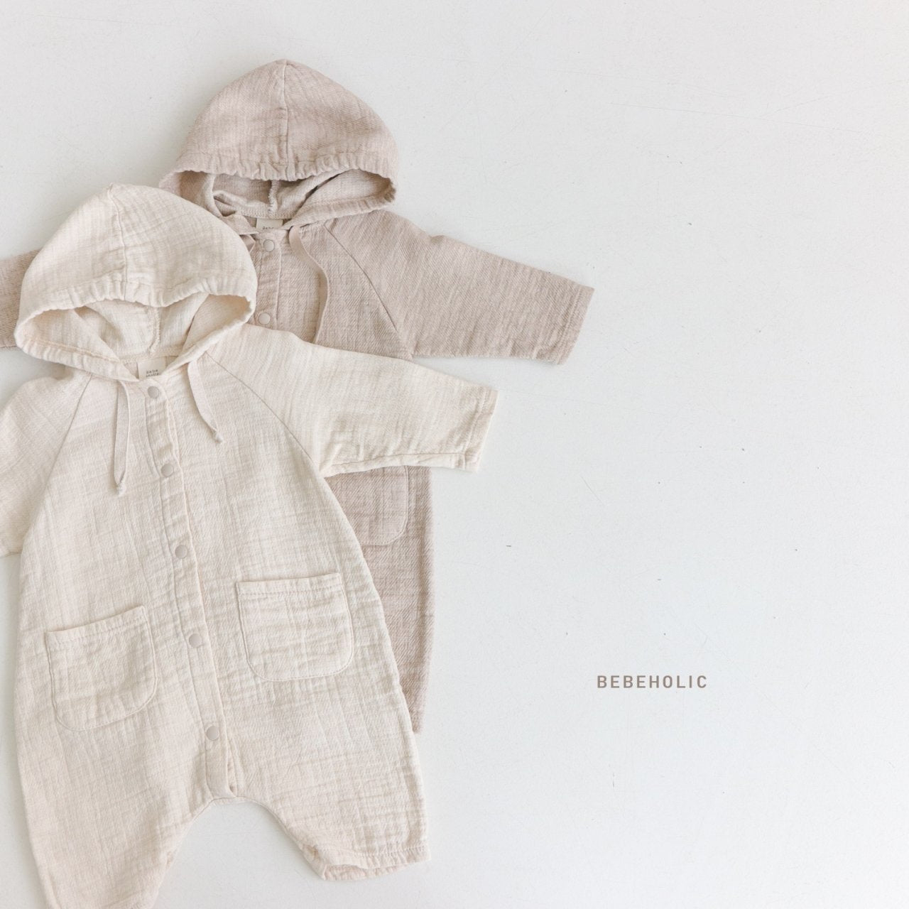 Zwei Baby-Strampler mit Kapuze, oder Hoody Bodysuits, liegen flach auf einem weißen Hintergrund. Der linke Strampler ist cremefarben und hat Vordertaschen, der rechte ist taupe. Unter den Hoody Bodysuits ist in Kleinbuchstaben der Text „Bebe Holic“ aufgedruckt.