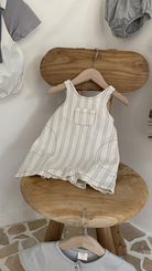 Ein ärmelloser, gestreifter Ariel St Body Suit von Bebe Holic mit kleiner Fronttasche hängt auf einem hölzernen Kleiderbügel, drapiert über einem rustikalen Holzstuhl. Weitere Babykleidung in verschiedenen Naturtönen und Karomuster hängen sichtbar in der Nähe. Der Boden ist mit Stein gefliest.