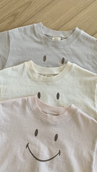 Drei gefaltete Bebe Holic Smile Tees aus weicher Baumwolle liegen gestapelt auf einer Holzfläche. Von oben nach unten sind die Farben hellgrau, cremefarben und hellrosa.