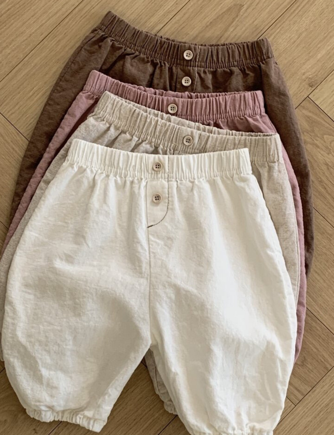 Vier Paar Natural Pants für Kinder von Bebe Holic liegen ordentlich gestapelt auf einem Holzboden. Diese vielseitigen Must-have-Shorts sind in den Farben Braun, Rosa, Beige und Weiß erhältlich und verfügen jeweils über eine elastische Taille und einen Knopf in der Mitte vorne.