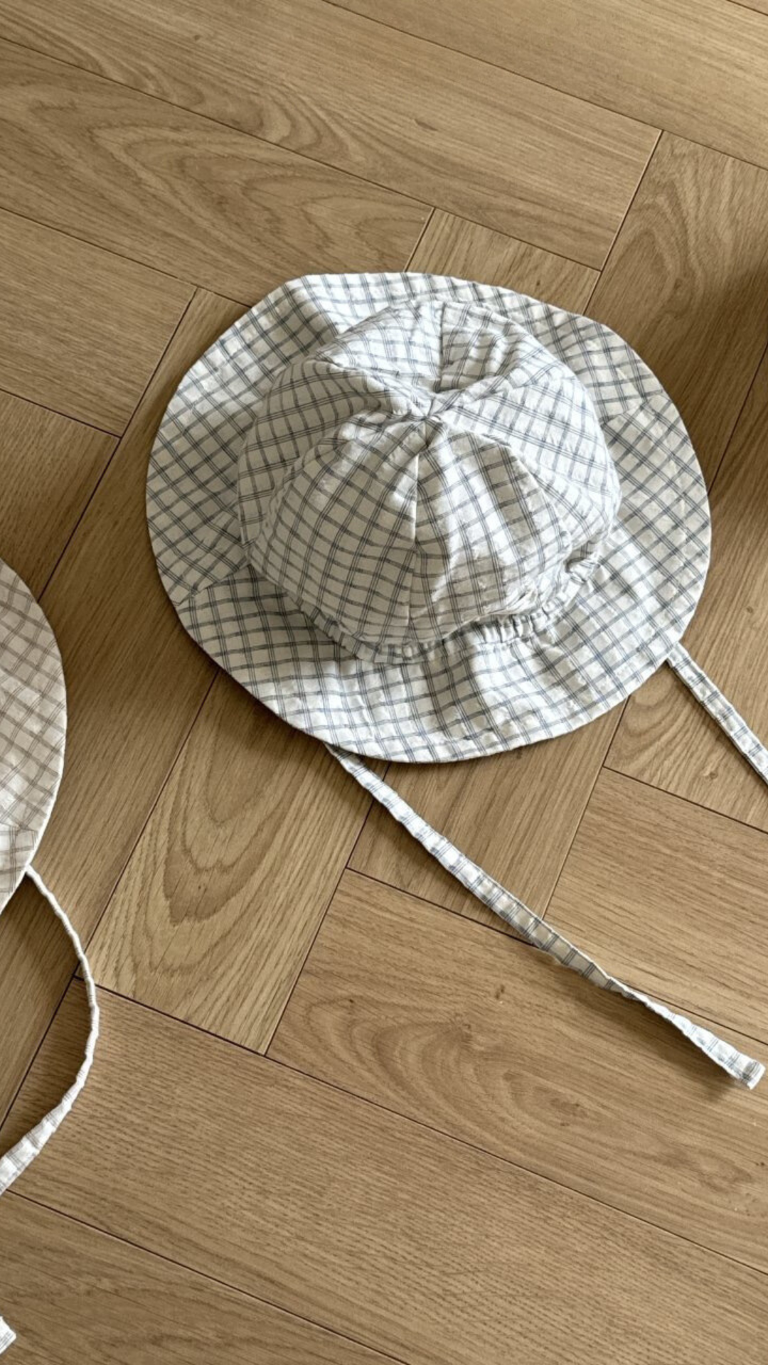 Ein weiß-blau karierter Yogurt ST Bucket Hat von Bebe Holic aus atmungsaktiven Materialien mit Kinnriemen liegt auf einem Holzboden mit Fischgrätenmuster. Ein weiterer ähnlicher Karo-Kinderhut ist teilweise in der Ecke sichtbar und bietet stylischen Sonnenschutz für junge Abenteurer.