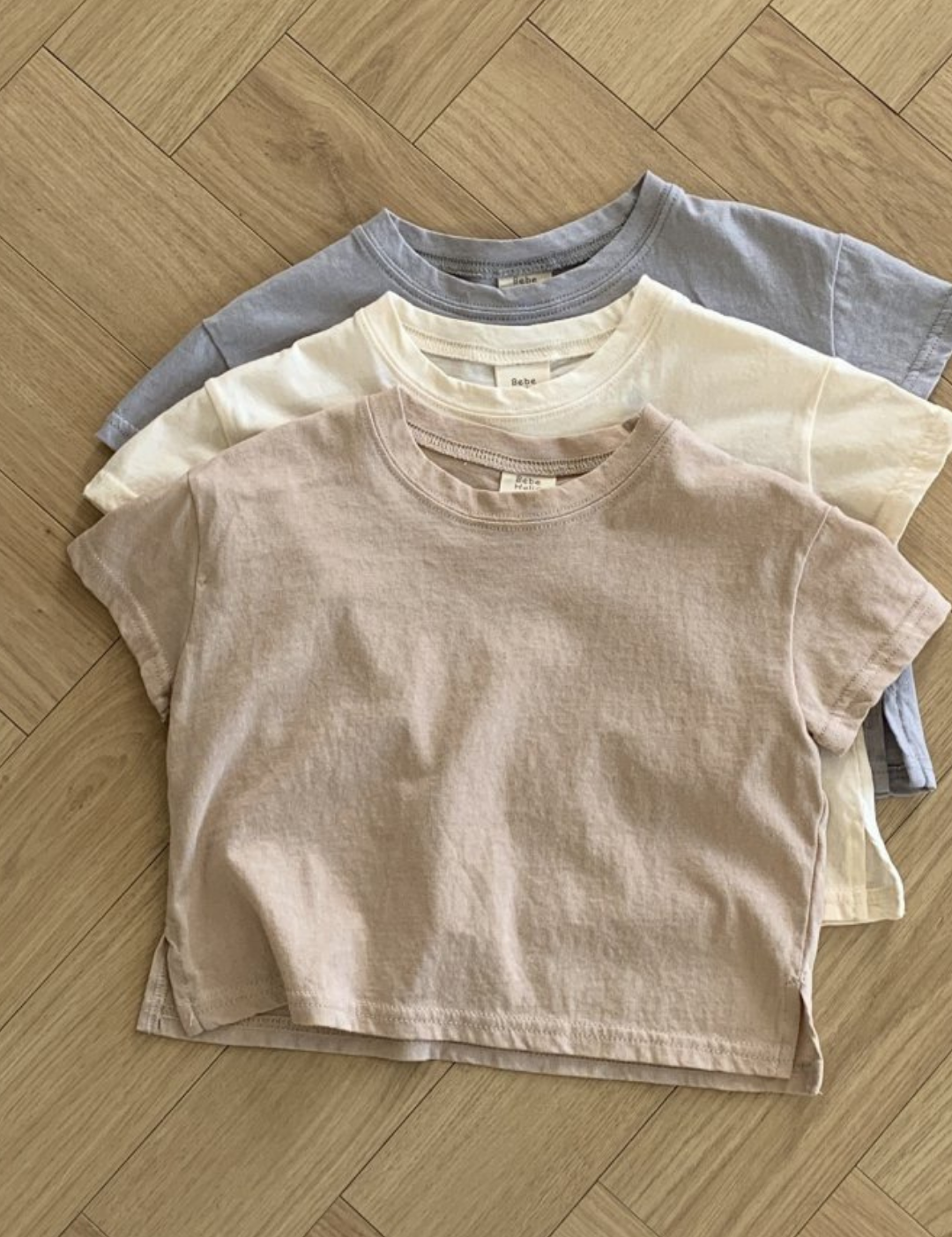 Drei pastellfarbene Basic-T-Shirts von Bebe Holic in Beige-, Creme- und Hellgrautönen sind ordentlich auf einem hölzernen Boden mit Fischgrätenmuster gestapelt. Diese T-Shirts aus hochwertiger Baumwolle haben eine lässige, entspannte Passform und kurze Ärmel in natürlichen Tönen.