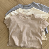 Drei pastellfarbene Basic-T-Shirts von Bebe Holic in Beige-, Creme- und Hellgrautönen sind ordentlich auf einem hölzernen Boden mit Fischgrätenmuster gestapelt. Diese T-Shirts aus hochwertiger Baumwolle haben eine lässige, entspannte Passform und kurze Ärmel in natürlichen Tönen.