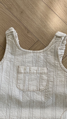 Ein cremefarbenes, strukturiertes Ariel St Body Suit Tanktop mit einer sichtbaren Vordertasche, flach auf einem Holzbodenhintergrund in Naturtönen von Bebe Holic gelegt.