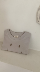 Ein ordentlich gefaltetes hellgraues T-Shirt aus weicher Baumwolle mit einem einfachen, braunen Smiley-Design auf der Vorderseite liegt auf einem weißen Regal vor einer hellen Wand. Auf der rechten Seite des Bildes hängt ein kleines Stück gemusterter Stoff, auf dem das Bebe Holic Smile Tee zu sehen ist.