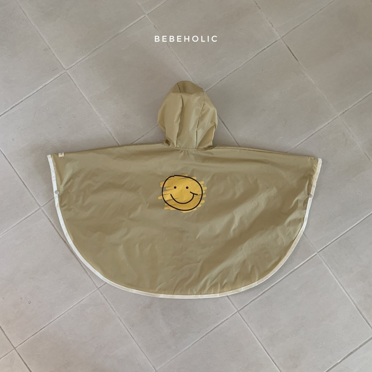 Ein beiger Smiley-Regenmantel 6m-24m - Der fröhliche Regenponcho für kleine Entdecker mit Kapuze liegt flach auf einem hellen Fliesenboden. Der wasserdichte Poncho hat in der Mitte ein einfaches, lächelndes gelbes Gesichtsemblem. Oben auf dem Bild ist das Wort „Bebe Holic“ aufgedruckt.