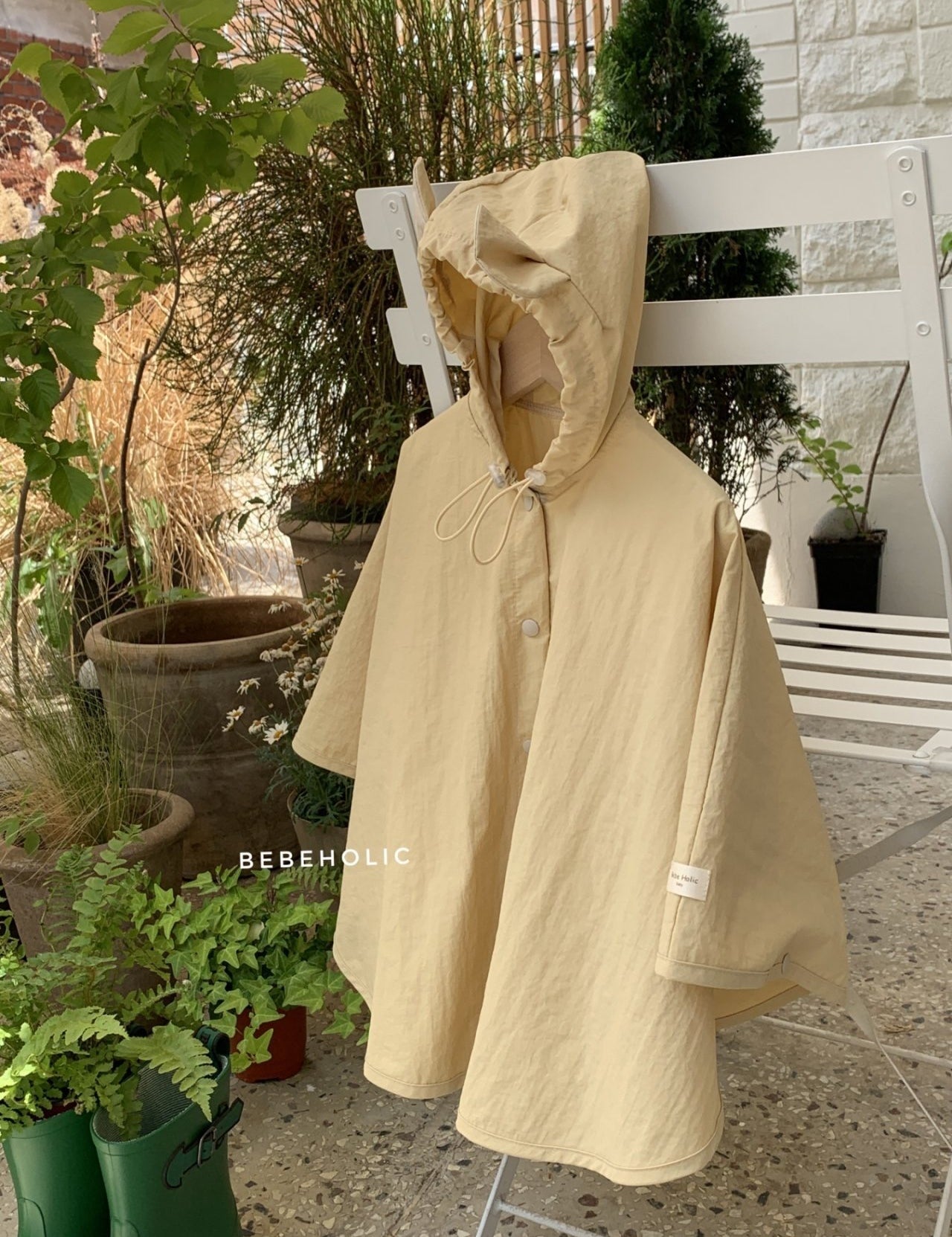 Ein beigefarbener Tierregenmantel mit Kapuze 6 m–24 m – Der perfekte Regenponcho für kleine Abenteurer von Bebe Holic steht auf einem weißen Stuhl in einer Außenumgebung, umgeben von Töpfen mit verschiedenen Pflanzen und Grünpflanzen. Der Poncho hat einen Knopf vorne, eine Kapuze mit Kordelzug und ist aus einem leichten wasserdichten Material gefertigt. Das Wort „BEBEHOLIC“ ist auf dem Stuhl sichtbar.