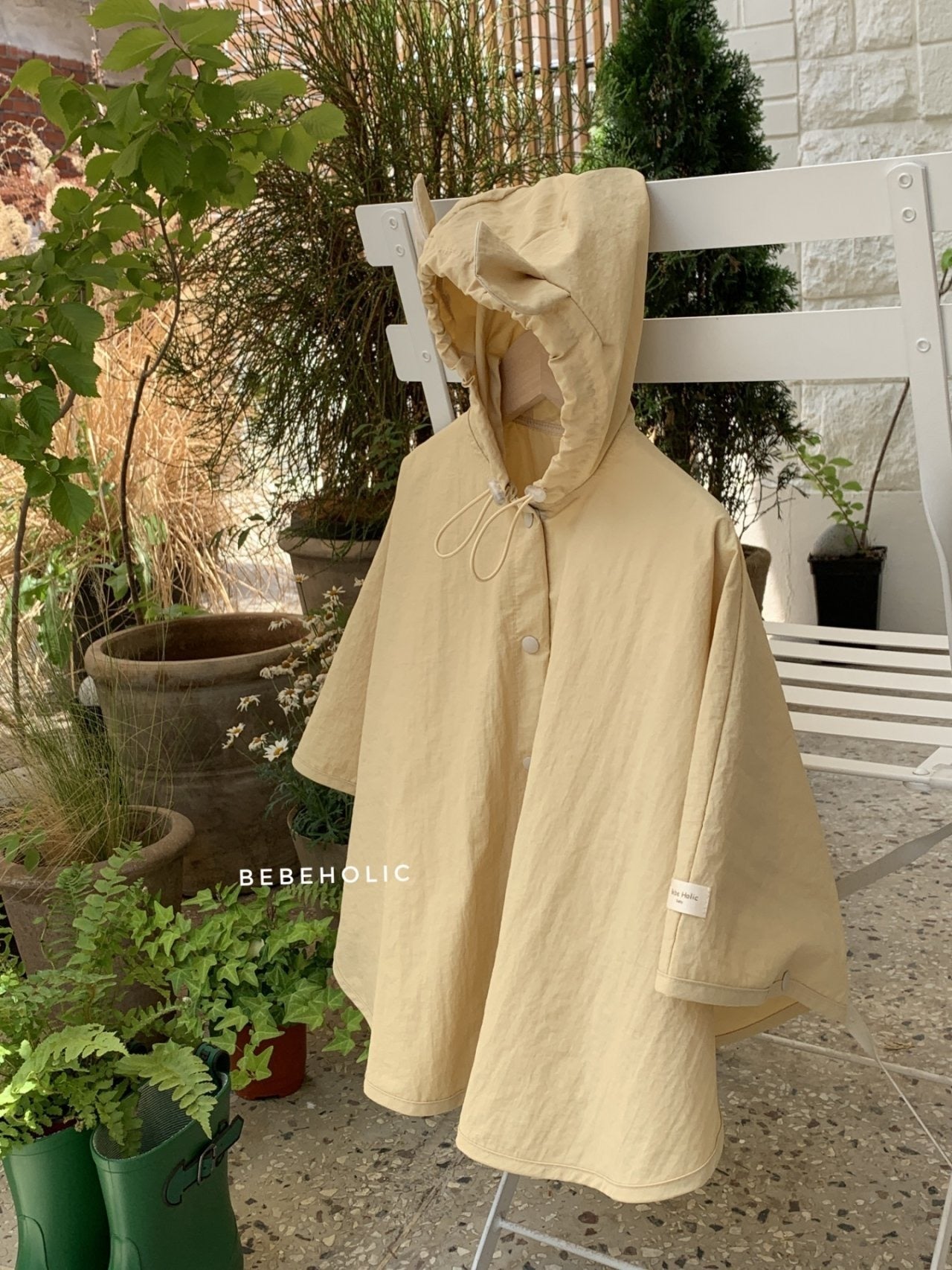 Ein beigefarbener Tierregenmantel mit Kapuze 6 m–24 m – Der perfekte Regenponcho für kleine Abenteurer von Bebe Holic steht auf einem weißen Stuhl in einer Außenumgebung, umgeben von Töpfen mit verschiedenen Pflanzen und Grünpflanzen. Der Poncho hat einen Knopf vorne, eine Kapuze mit Kordelzug und ist aus einem leichten wasserdichten Material gefertigt. Das Wort „BEBEHOLIC“ ist auf dem Stuhl sichtbar.