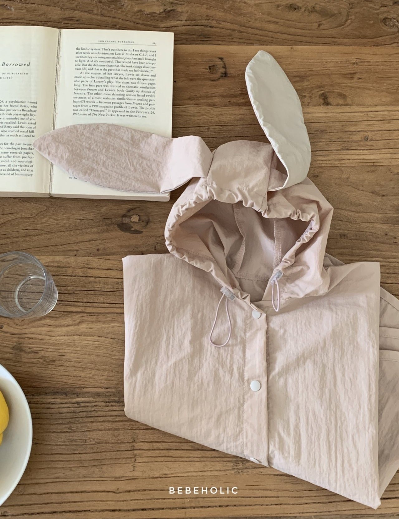 Ein rosa **Tierregenmantel 6m-24m - Der perfekte Regenponcho für kleine Abenteurer** mit Hasenohrenkapuze liegt zusammengefaltet auf einem Holztisch neben einem offenen Buch. Ein durchsichtiges Trinkglas und ein Teller mit ein paar gelben Früchten stehen ebenfalls auf dem Tisch. Der Text „BEBEHOLIC“ ist unten im Bild zu sehen und unterstreicht seine bequeme Passform.