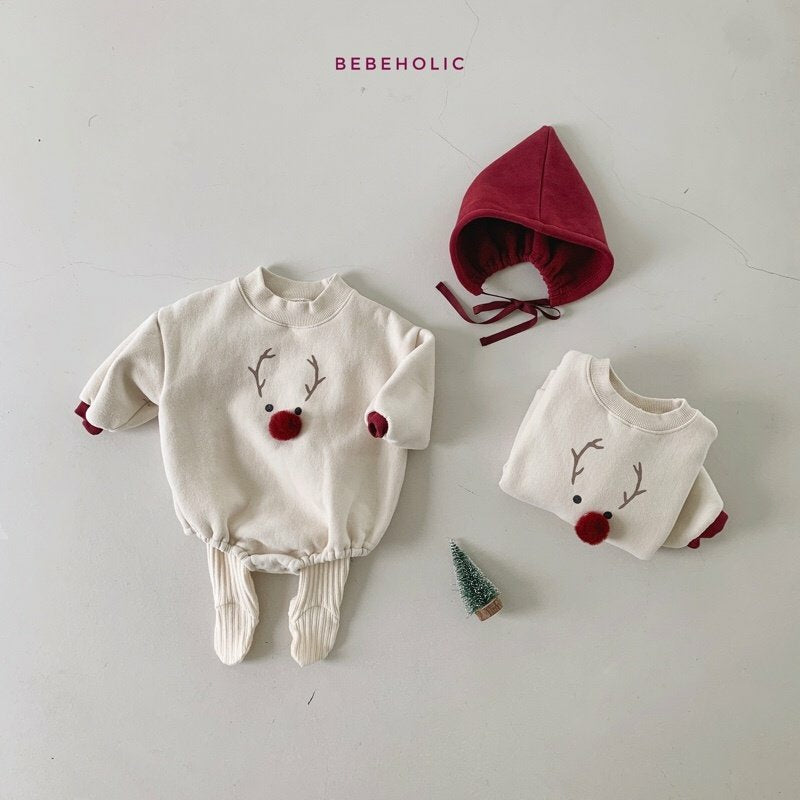 Zwei weiße Baby-Outfits mit Rentiermotiven und roten Bommelnasen liegen auf einer hellen Unterlage. Ein Outfit hat lange Hosen und das andere ist gefaltet. Eine rote Kapuze mit Bändern und ein Miniatur-Weihnachtsbaum vervollständigen diesen festlichen Look. „Bebe Holic“ steht darüber, was dieses Hirschhauben-Set kuschelig und entzückend macht.