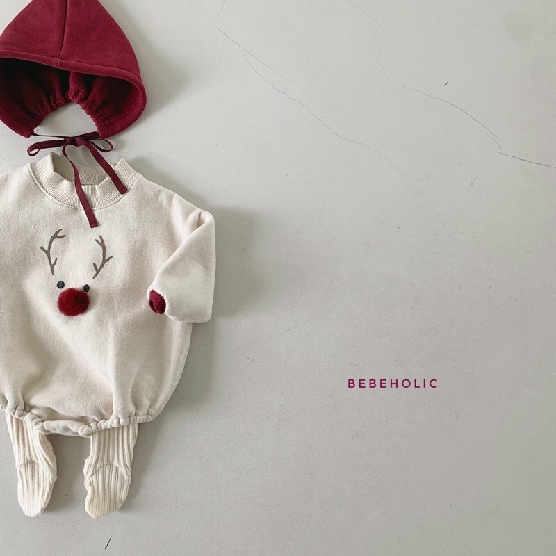 Ein Babyoutfit wird flach auf eine helle Oberfläche gelegt. Der cremefarbene Strampler zeigt ein Rentiergesicht mit Geweih und einer roten Bommelnase, ergänzt durch eine festliche Kapuze mit braunen Bändern. Der Markenname Bebe Holic ist auf der rechten Seite in Pink aufgedruckt, perfekt für einen kuscheligen und entzückenden Look.