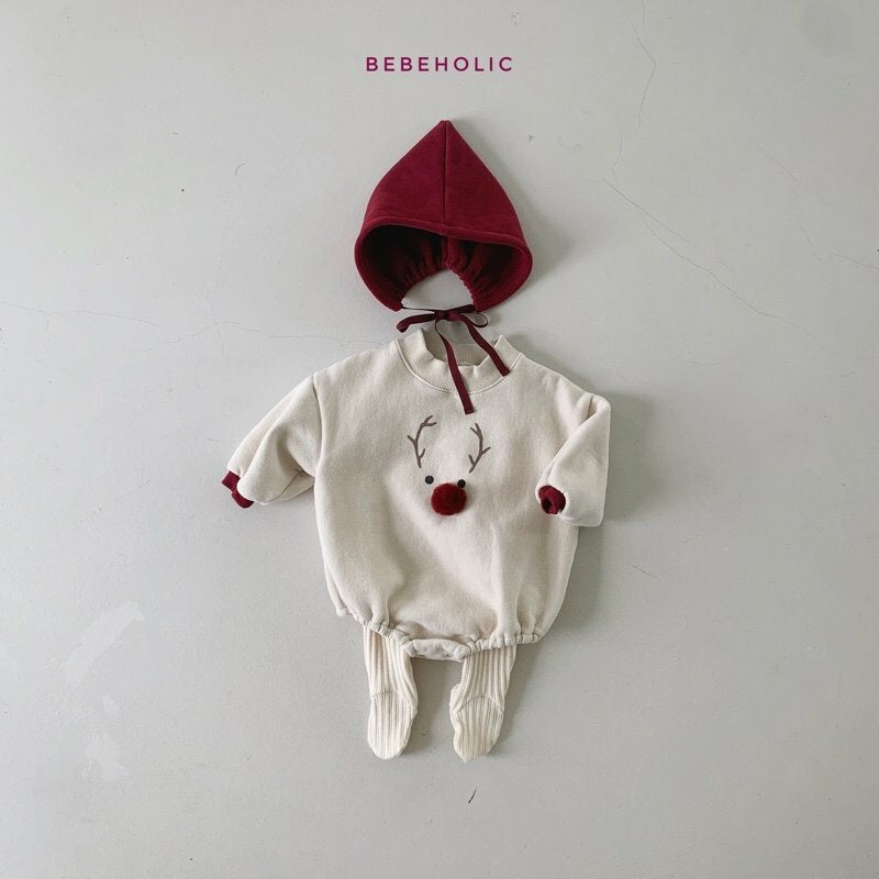 Ein Baby-Outfit wird auf einer flachen Oberfläche präsentiert. Das Set besteht aus einem beigen Einteiler mit einem Rentiergesicht und Geweih, das auf der Vorderseite aufgestickt ist, mit einer roten Nase und passenden roten Manschetten. Eine kastanienbraune Kapuze mit Bändern und weiße Leggings mit Füßen vervollständigen das Ensemble, perfekt für einen festlichen Look. Oben ist der Schriftzug „Bebe Holic Deer Bonnet Set“ aufgedruckt.