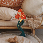 Eine skurrile Little Lights Ariel Mermaid Lampe mit roten Haaren und grünem Schwanz, die auf einem Nachttisch aus Bambus mit flauschigen Kissen und Rattan-Accessoires in einem gemütlich eingerichteten Zimmer steht.