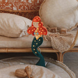 Eine skurrile Little Lights Ariel Mermaid Lampe mit roten Haaren und grünem Schwanz, die auf einem Nachttisch aus Bambus mit flauschigen Kissen und Rattan-Accessoires in einem gemütlich eingerichteten Zimmer steht.