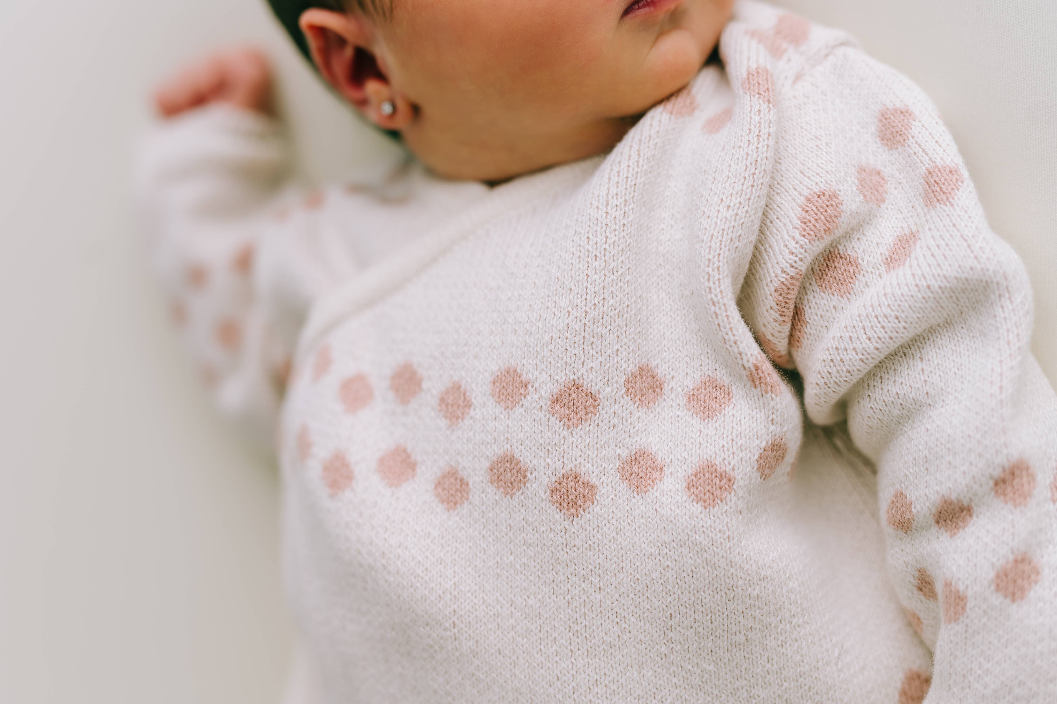 Nahaufnahme eines Babys, das einen beigen Strickpullover von Micu Micu aus dem Newborn Pack Punkte rosa: 0 -3 Monat (56 cm) trägt und rosa geometrische Formen um den Brustbereich aufweist. Das Baby hat kleine Ohrringe und liegt, wobei ein Teil seines Gesichts und Oberkörpers sichtbar ist.